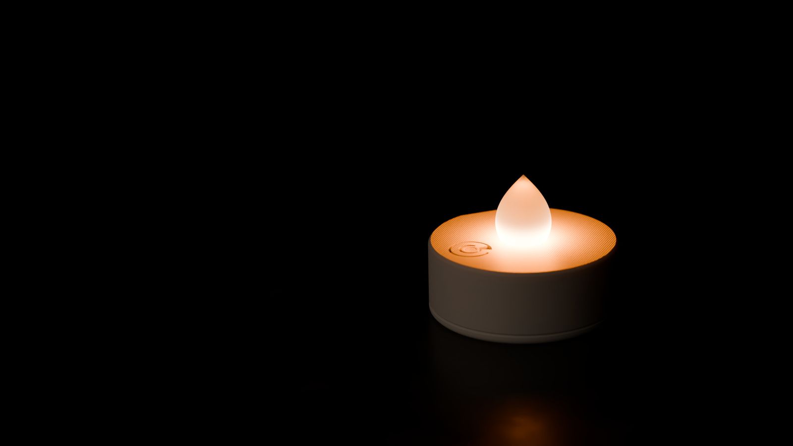 A glowing teelicht pro on dark stone: a short ivory cup with a frosted teardrop on top, lit from inside.